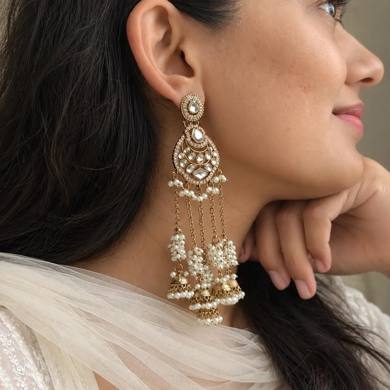 A woman in an ivory dupatta, seen in profile, wearing a long gold-tone kundan chandbali with pearl clusters and small jhumka drops